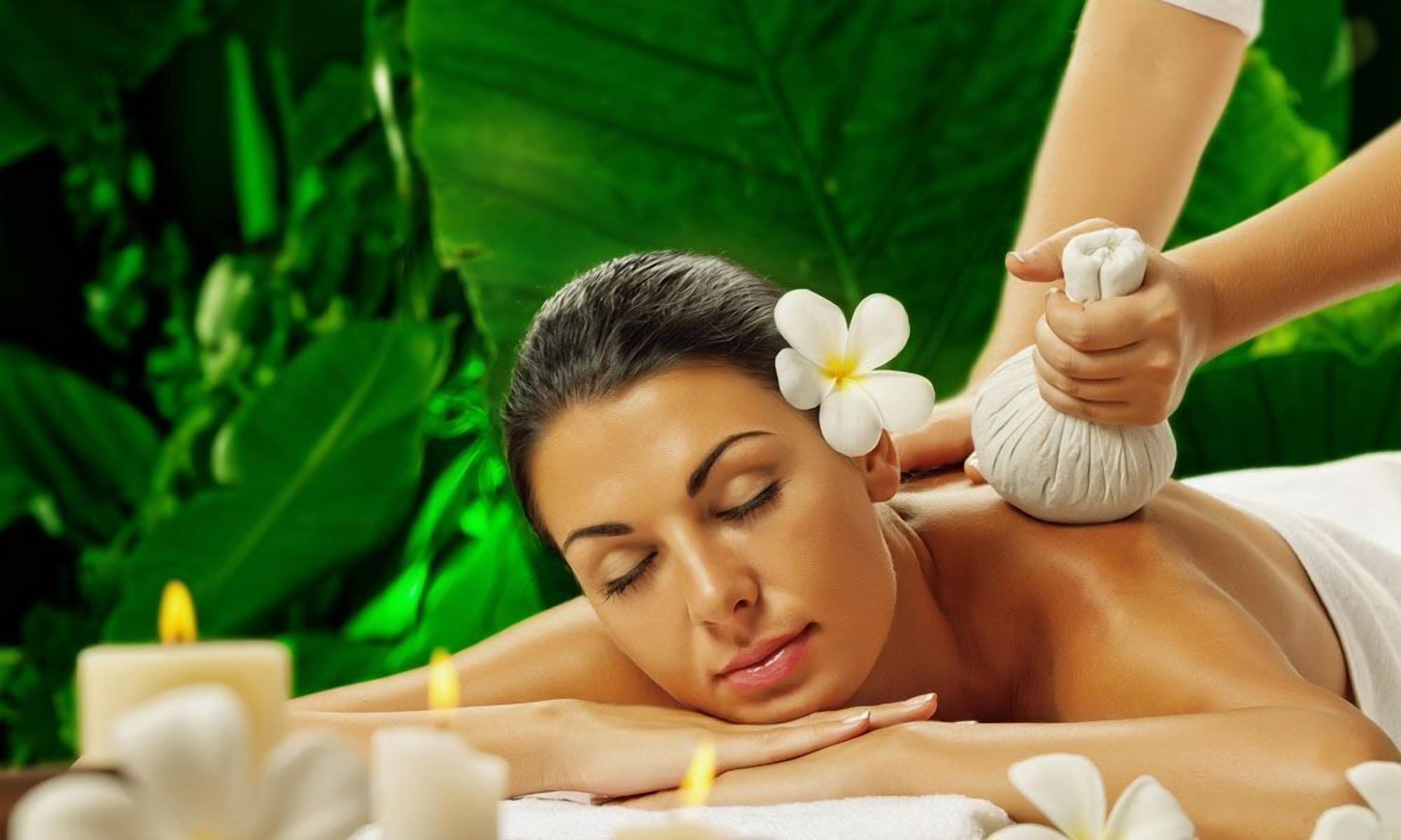 A person receiving a massage with a flower in their hair, surrounded by candles and greenery.