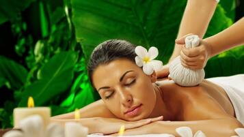 A person receiving a massage with a flower in their hair, surrounded by candles and greenery.