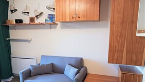 Una habitación pequeña y acogedora con un sofá gris, armarios de madera y una estantería con diversos objetos, incluyendo un dardo y libros.