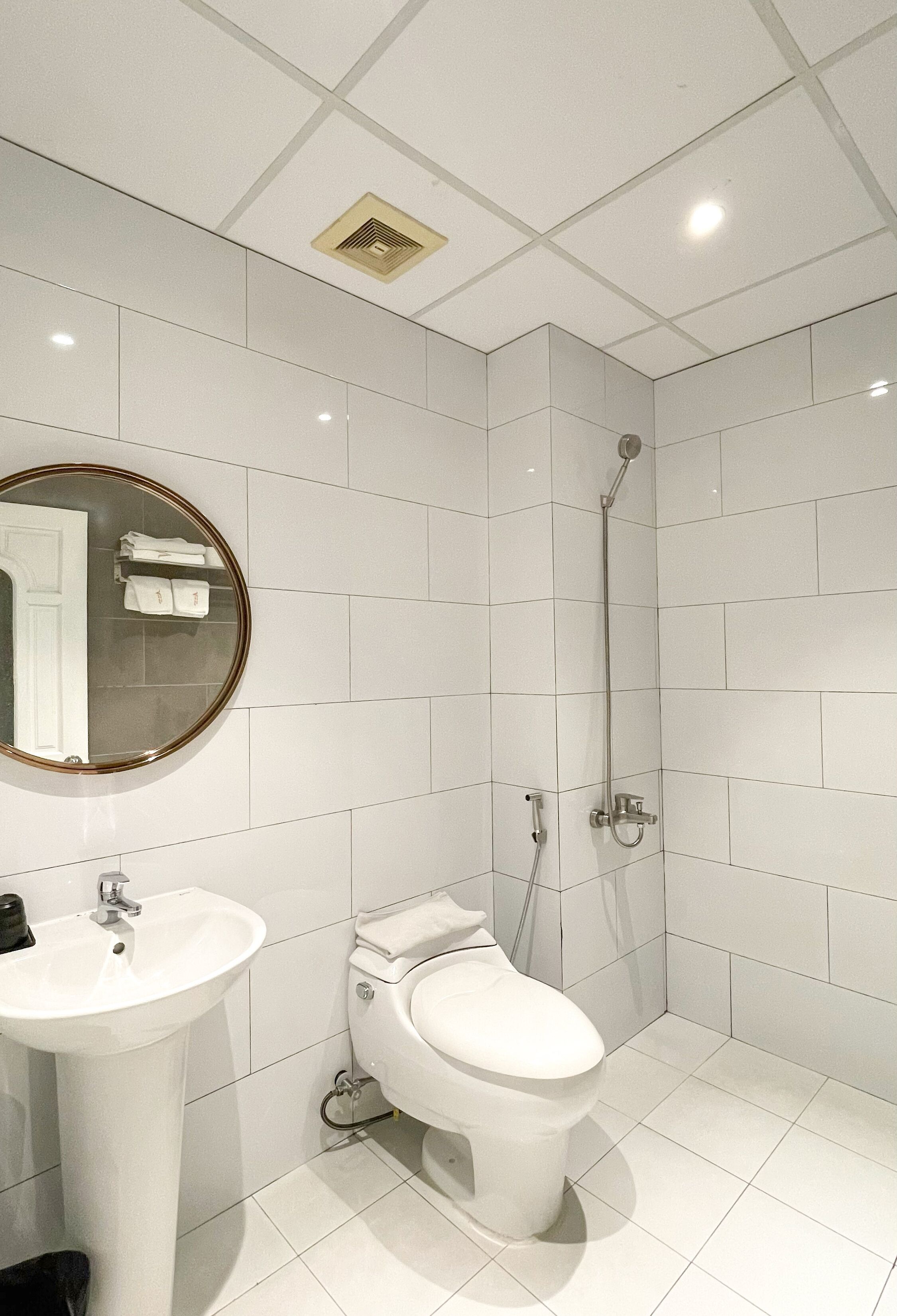 Ein modernes Badezimmer mit weißen Fliesen, einem runden Spiegel, einem Waschbecken, einer Toilette und einer Dusche.
