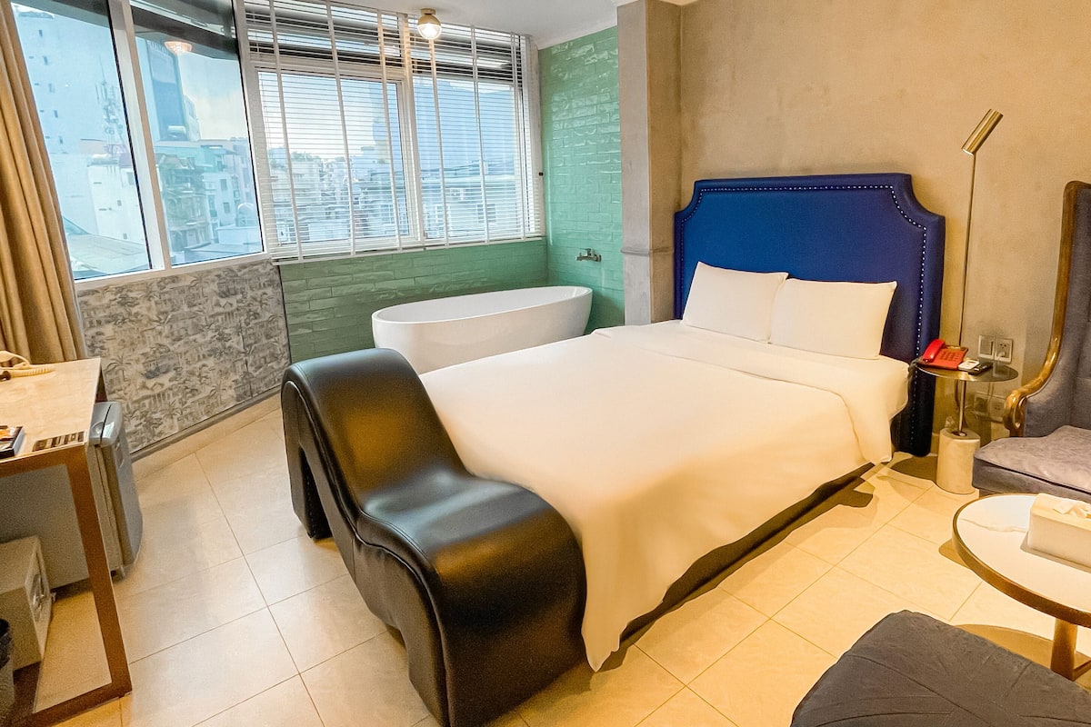 Une chambre d’hôtel comprenant un lit, un bureau, une chaise, une petite table, un téléphone et une fenêtre avec des stores.