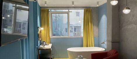 Una habitación de hotel con cama, una mesita redonda pequeña, una silla, un lavamanos y una ventana con cortinas.