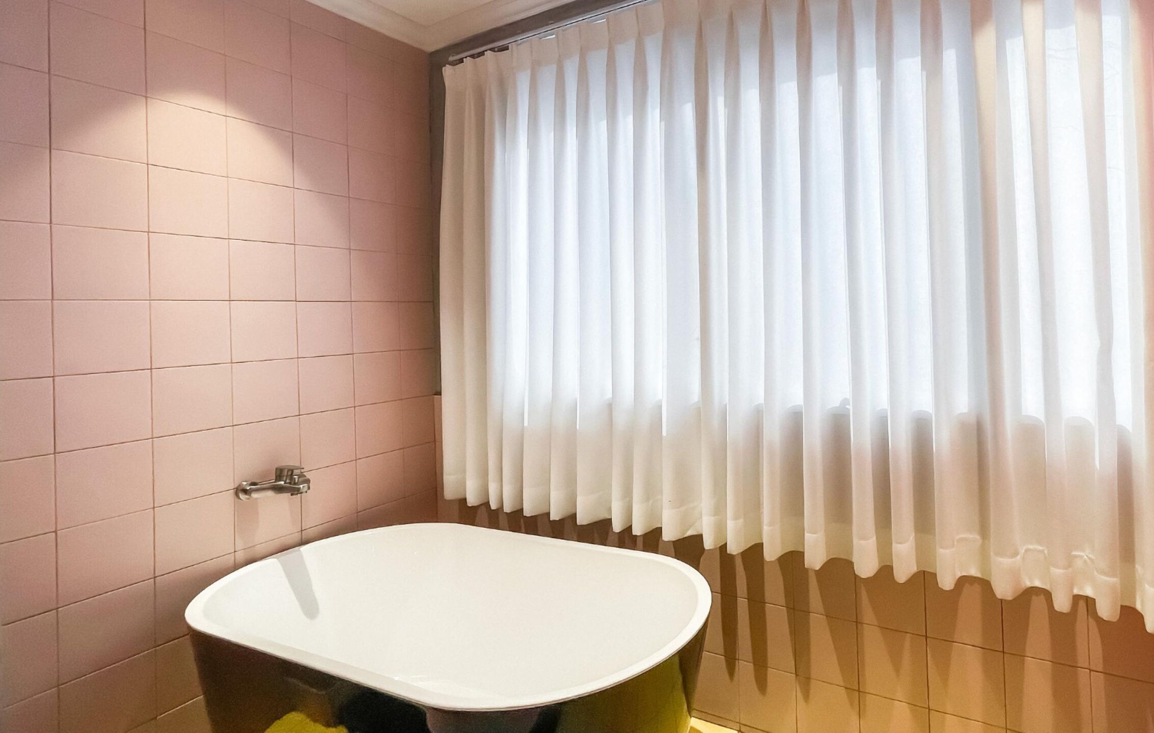 Un baño con paredes de azulejos rosas, una bañera blanca y una ventana con cortinas blancas.