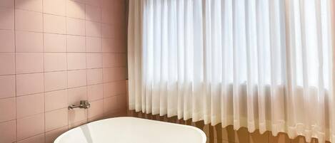A bathroom with pink tiled walls, a white bathtub, and a window with white curtains.