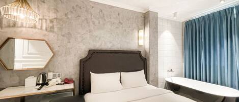 A hotel room with a bed, bedside table, lamp, mirror, and a bathroom with a bathtub and curtains.