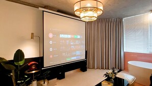 A modern living room with a large flat-screen TV, a dining table with a vase of flowers, a potted plant, and a view of a red brick wall through the window. - A IN HOTEL DEL LUNA (Ho Chi Minh City)