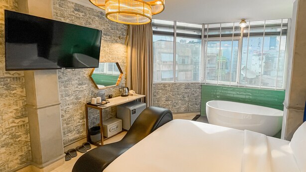 A hotel room with a flat-screen TV mounted on the wall, a desk with a computer, a chair, a large bed, a bathtub, and a window with blinds. - A IN HOTEL DEL LUNA (Ho Chi Minh City)