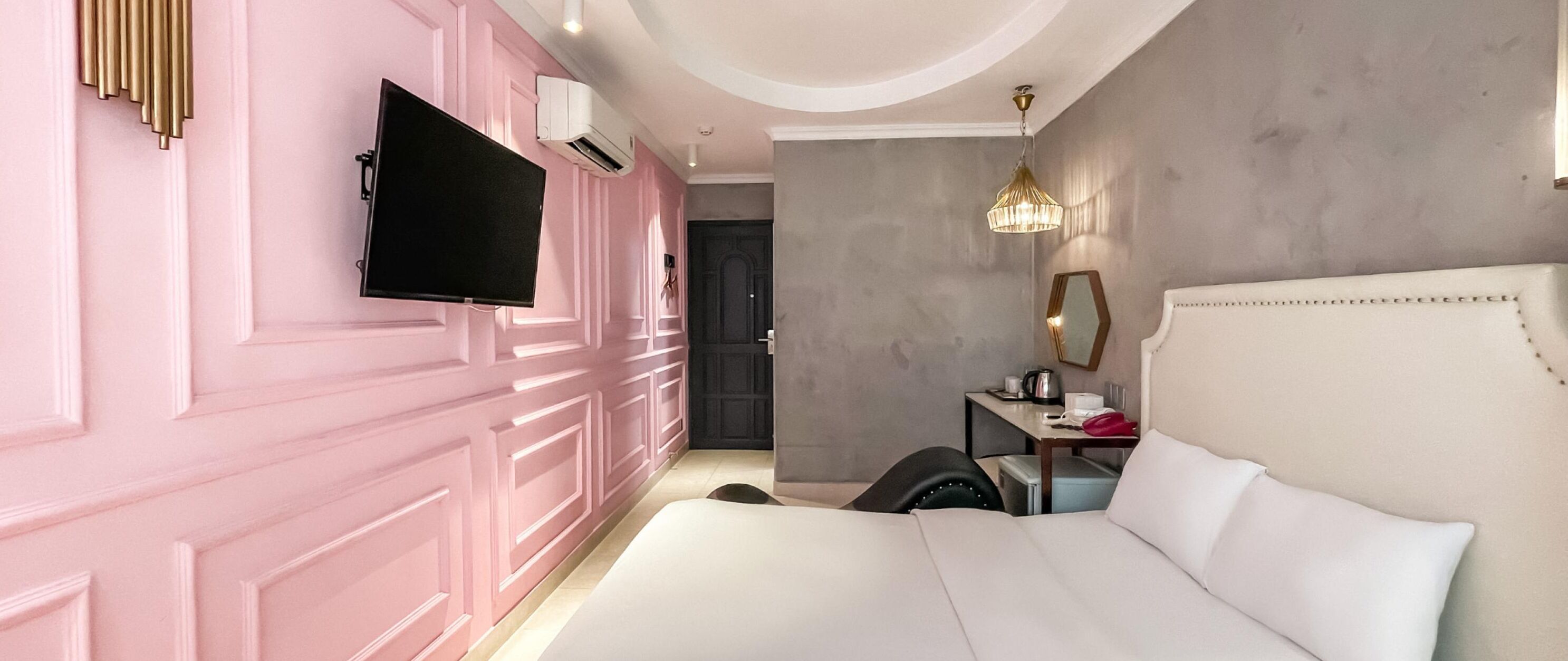 Una habitación de hotel moderna con una pared rosa, un televisor de pantalla plana, una cama con ropa blanca, un pequeño escritorio con una lámpara y una pared gris con textura.