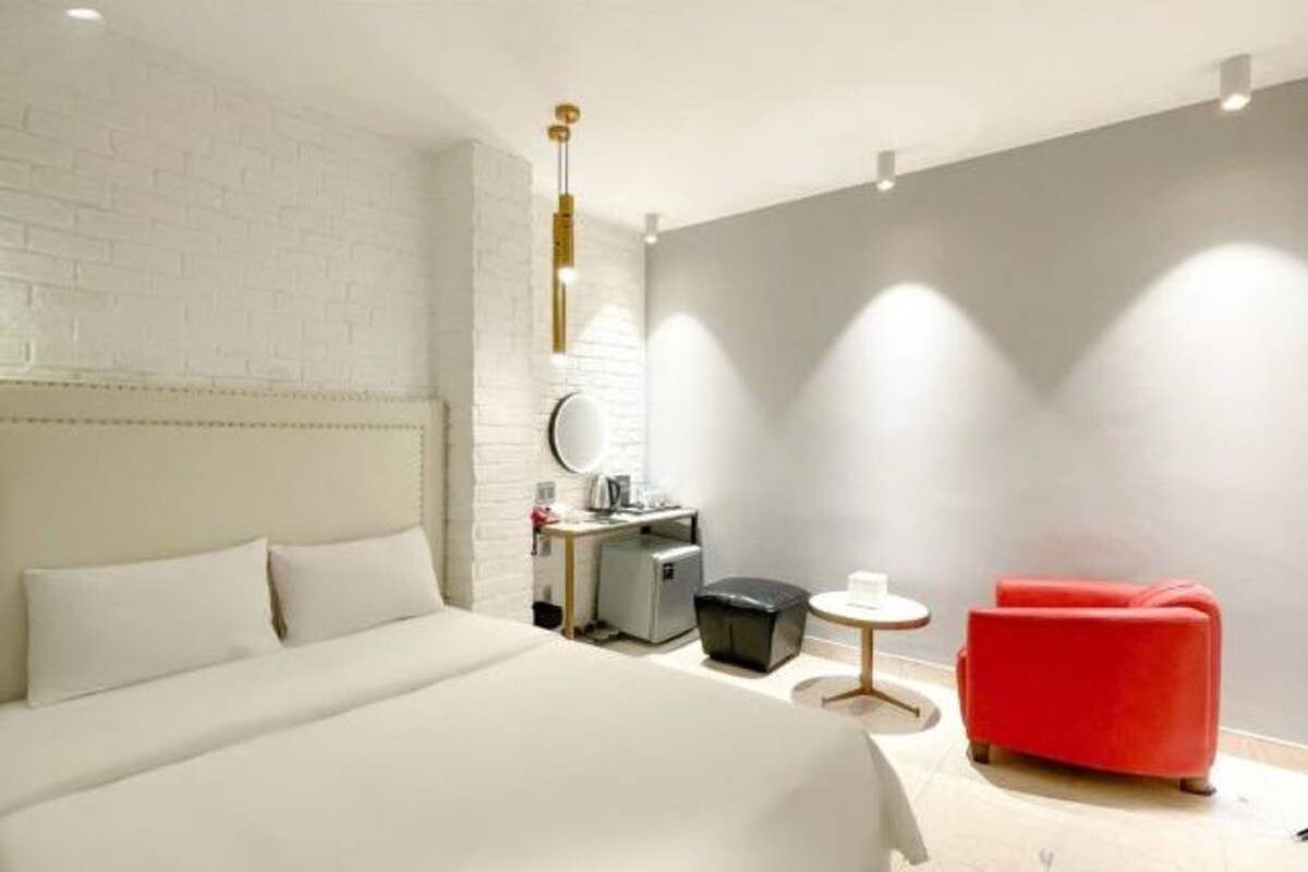 Ein modernes Hotelzimmer mit einem großen Bett, einem roten Schlafsofa, einem kleinen Schreibtisch mit Spiegel und einer kompakten Küchenzeile.
