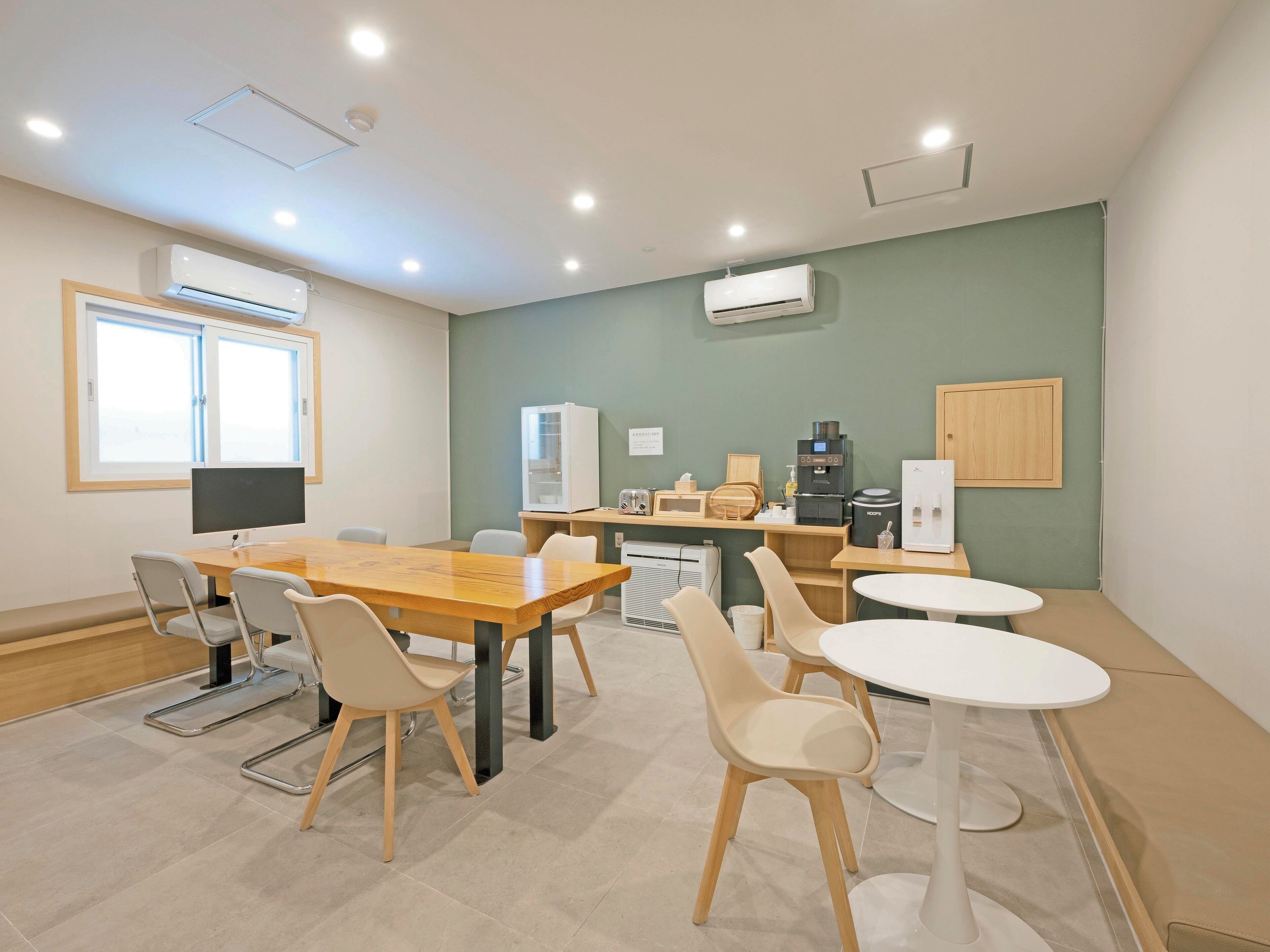 A modern, well-lit room with a wooden table and chairs, a refrigerator, a coffee machine, and a microwave.