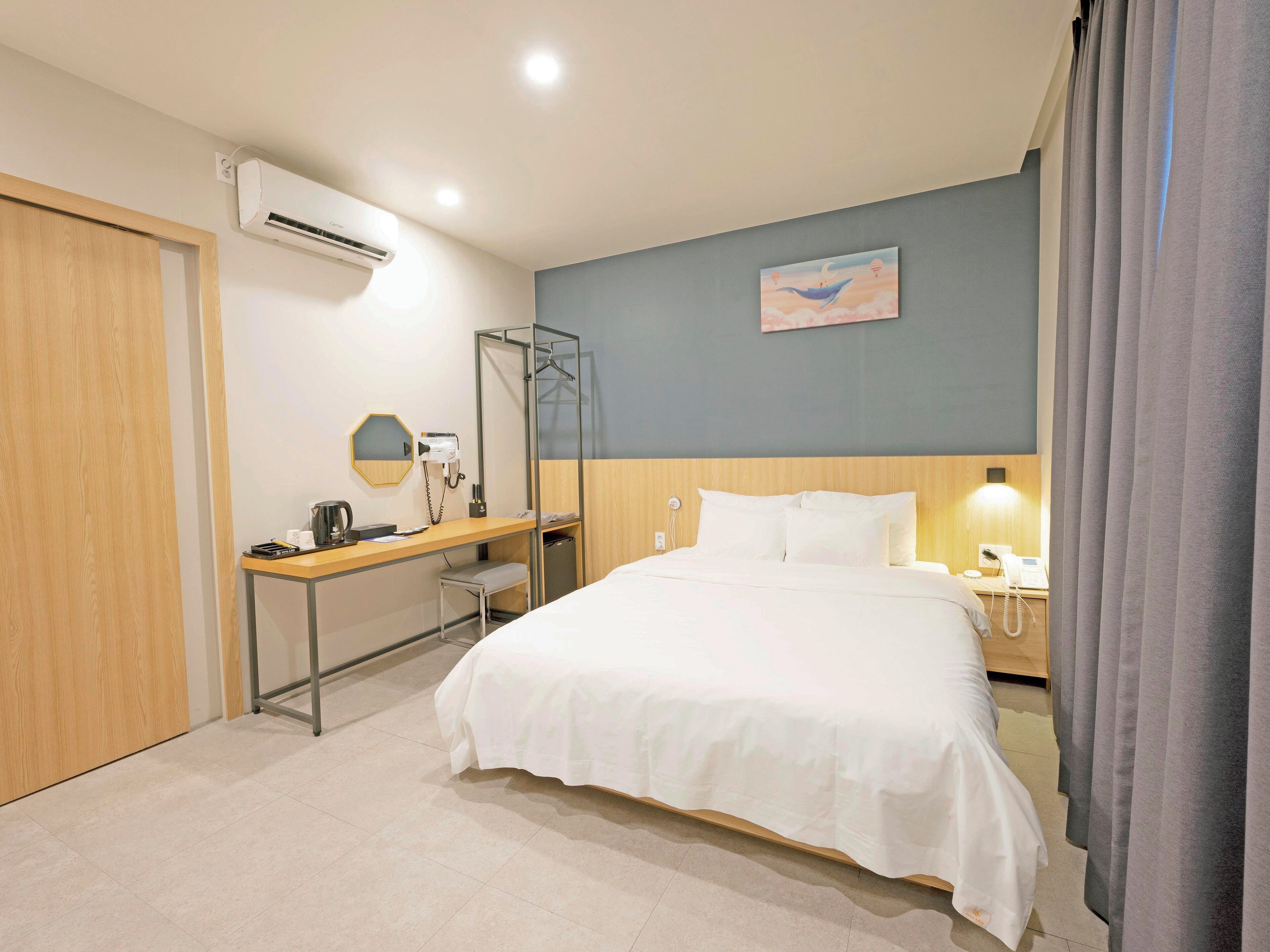 Quarto de hotel com uma cama, uma escrivaninha com cadeira, um espelho, um telefone e um quadro na parede.