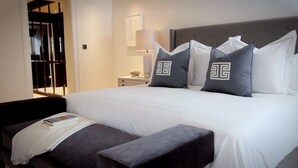 A neatly made bed with white bedding and grey pillows, a bedside table with a lamp, and a dark-colored bench at the foot of the bed. - Beautiful 4-bed Flat in Mayfair (London)