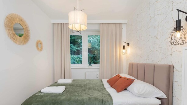 Un dormitorio ordenado con una cama, una ventana con cortinas, un espejo montado en la pared y dos apliques.