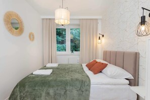 Un dormitorio ordenado con una cama, una ventana con cortinas, un espejo montado en la pared y dos apliques.