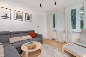 Un salon moderne avec un canapé gris, une table basse ronde et une vue sur une forêt, visible à travers de grandes fenêtres.