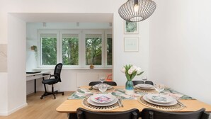 Un comedor con una mesa de madera dispuesta para dos personas, dos sillas, un florero con flores y una ventana con vistas a árboles.