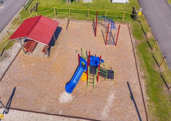 A playground with a blue slide and climbing structures, surrounded by a fenced area with a red-roofed building nearby.