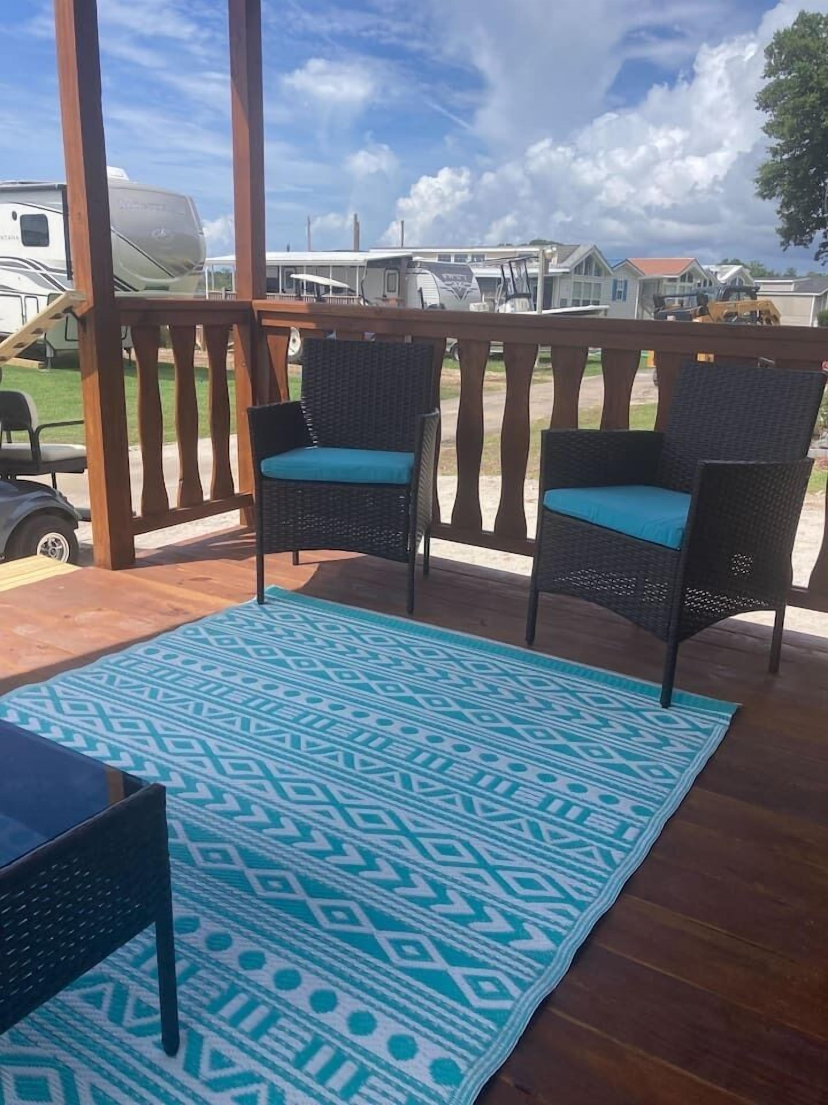 Ein überdachter Außenbereich mit zwei Korbstühlen, blauen Kissen, einem gemusterten blauen Teppich und einer Holzterrasse.