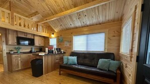 A cabin interior with a wooden floor, a sofa with cushions, a kitchen area with a microwave, and a window with blinds. - Family + Pet-Friendly Cabin w/ Loft | Surf City (Holly Ridge)
