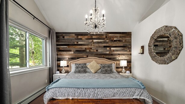 A bedroom with a wooden headboard, a chandelier, a bed with patterned bedding, bedside lamps, and a view of trees through a window. - Cozy Creek! End Unit with Creekside Patio, Grill, Top Deck & Communal Hot Tub! (Steamboat Springs)