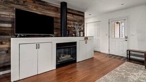 A modern living room with a fireplace, white cabinets, a large flat-screen TV, and a wooden accent wall. - Cozy Creek! End Unit with Creekside Patio, Grill, Top Deck & Communal Hot Tub! (Steamboat Springs)