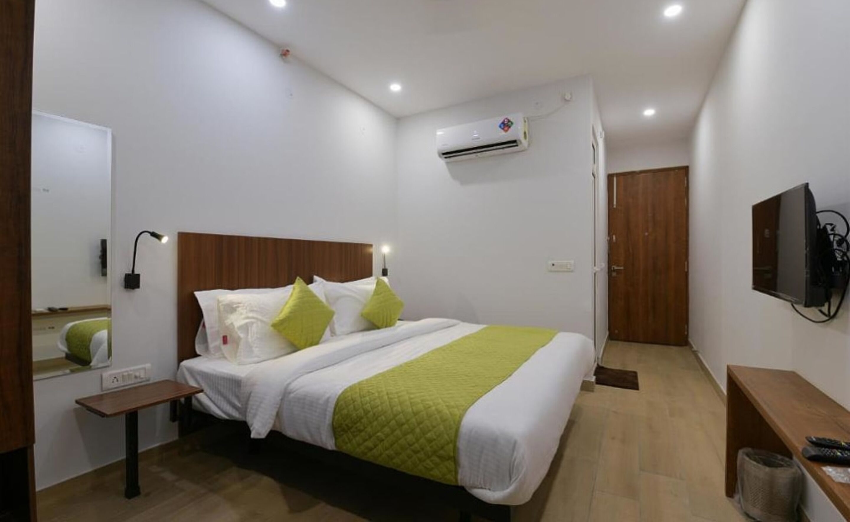 A hotel room with a bed, bedside table, wall-mounted TV, air conditioning unit, and a wooden door.