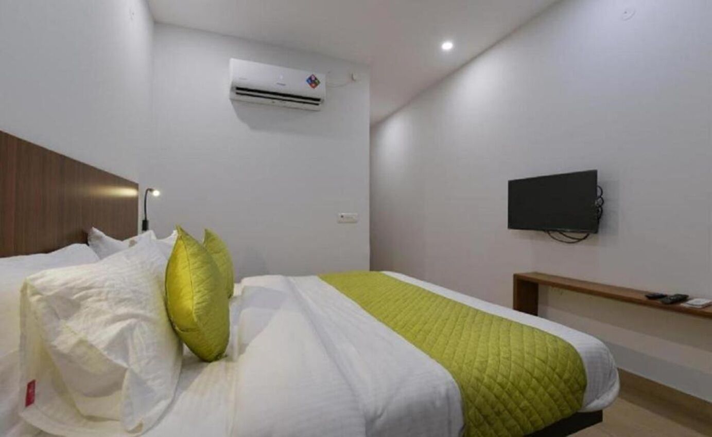 Ein Hotelzimmer mit Bett, Kissen, einer gesteppten grünen Decke, einer Klimaanlage, einem an der Wand montierten Flachbildfernseher und einem Nachttisch.