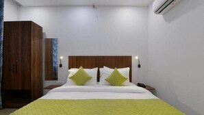 A hotel room with a wooden headboard, a bed with white bedding and green pillows, a nightstand with a lamp, and an air conditioning unit on the wall. - Hotel The Emporio A Corporate suites (Ghaziabad)