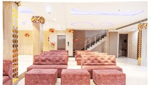 A spacious lobby with multiple pink tufted ottomans arranged in rows, a staircase, and a modern elevator. - Hotel The Emporio A Corporate suites (Ghaziabad)