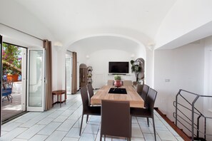 Ein geräumiger Essbereich mit Holztisch und modernen Stühlen, einem Wand-Fernseher und einer Glastür, die auf eine Außenpatio mit Pflanzen in Töpfen führt.