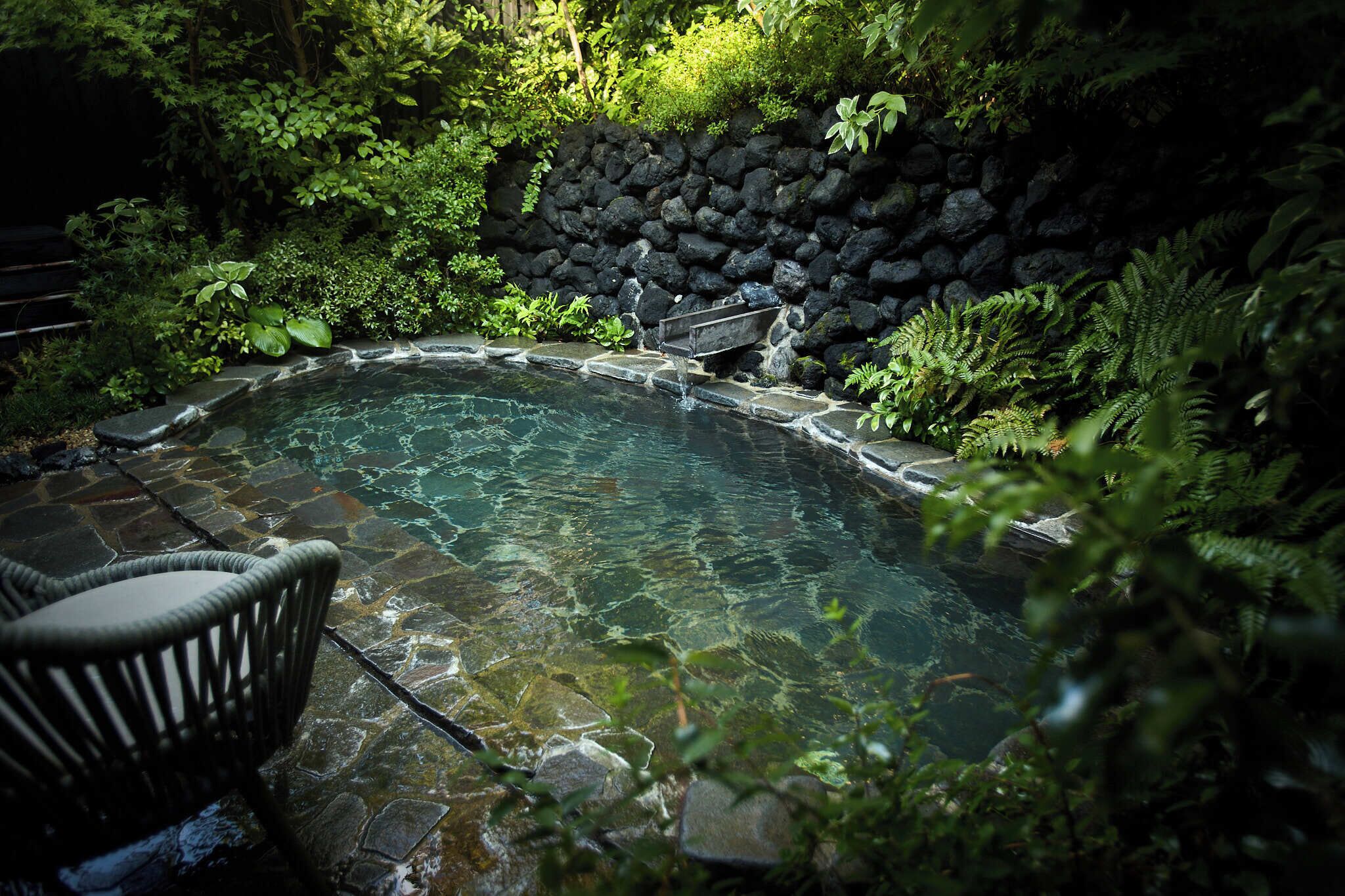 A stone hot tub surrounded by lush greenery and a stone wall.