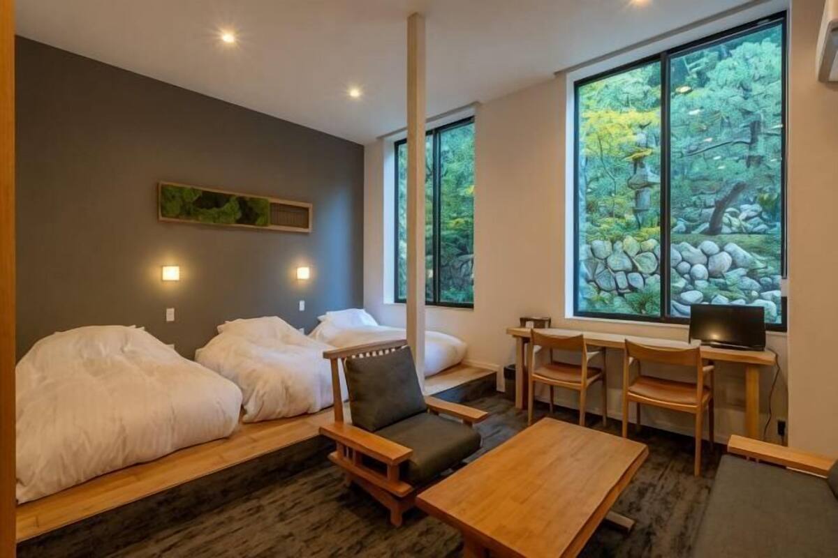 Ein Zimmer mit zwei Betten, einem Schreibtisch und Blick auf Bäume durch große Fenster.
