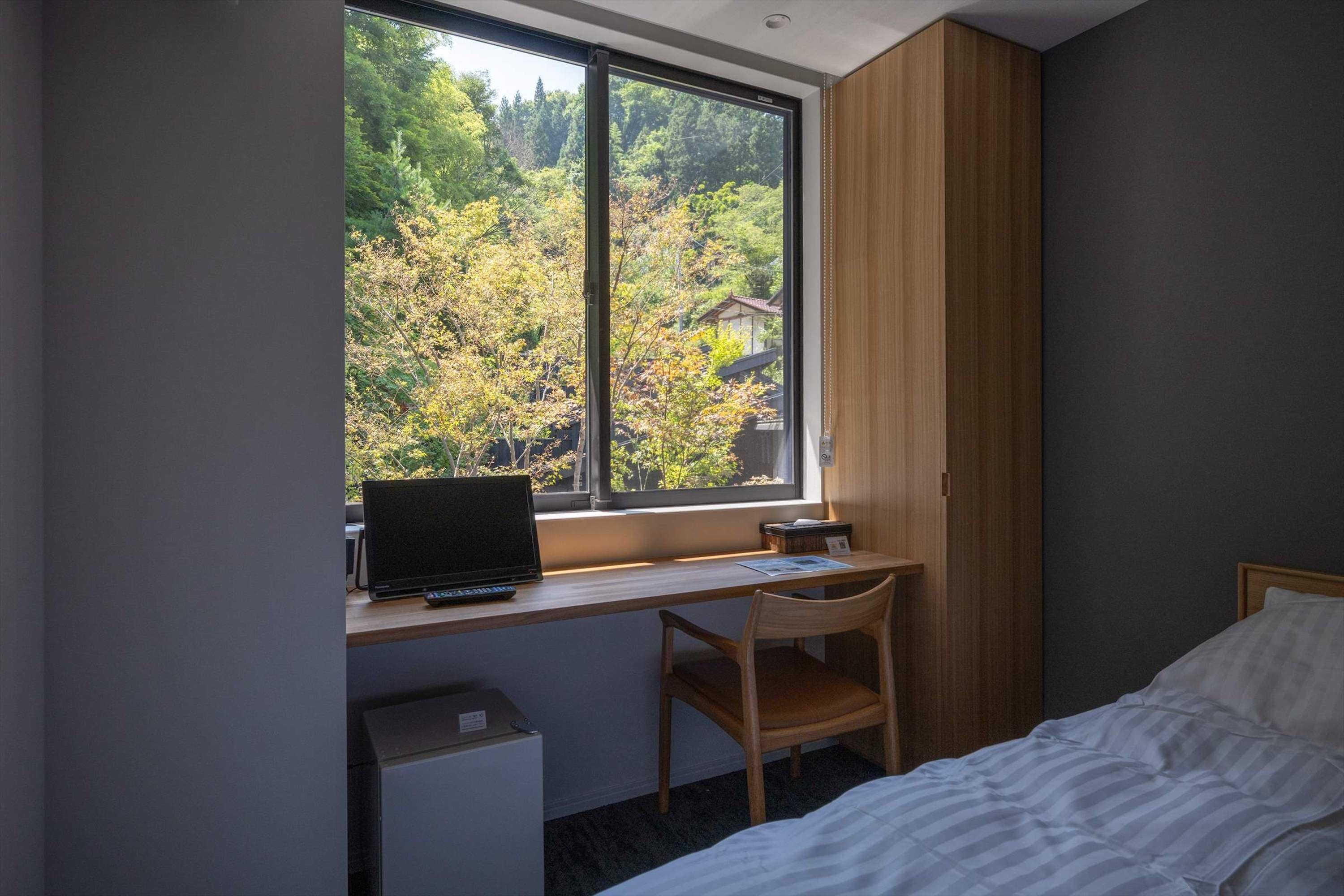 Une chambre à coucher avec un lit, un bureau avec un ordinateur portable, une chaise et une fenêtre donnant sur des arbres et une maison.