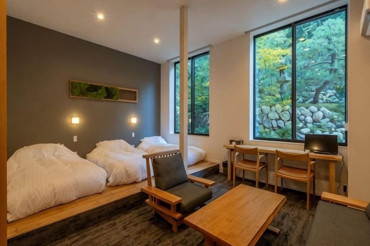 A room with two beds, a desk, and a view of trees through large windows.