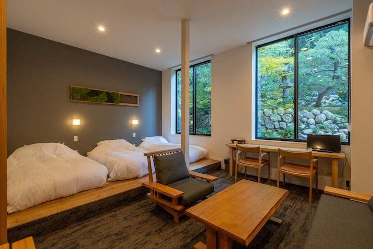 Ein Zimmer mit zwei Betten, einem Schreibtisch und einem Fernseher. Fenster bieten Blick auf Bäume und Felsen.