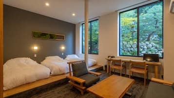 A room with two beds, a desk, and a TV. There are windows with a view of trees and rocks.