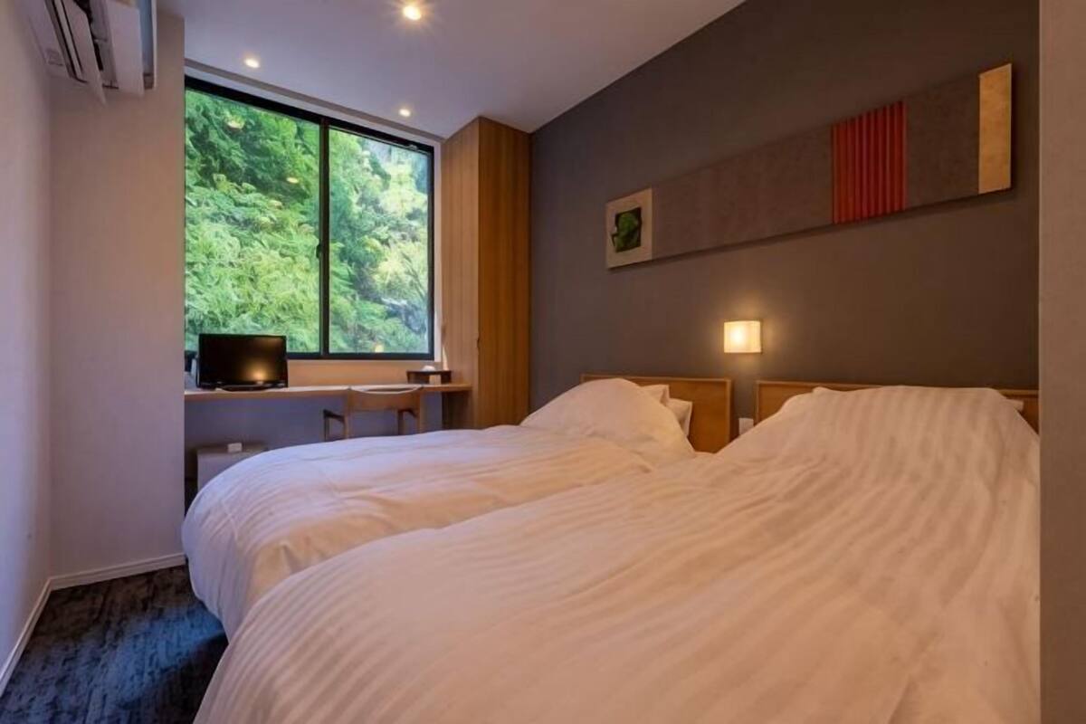 Ein Schlafzimmer mit einem großen Bett, einem Schreibtisch mit Stuhl, einem Fenster mit Blick ins Grüne und einer Wandleuchte.