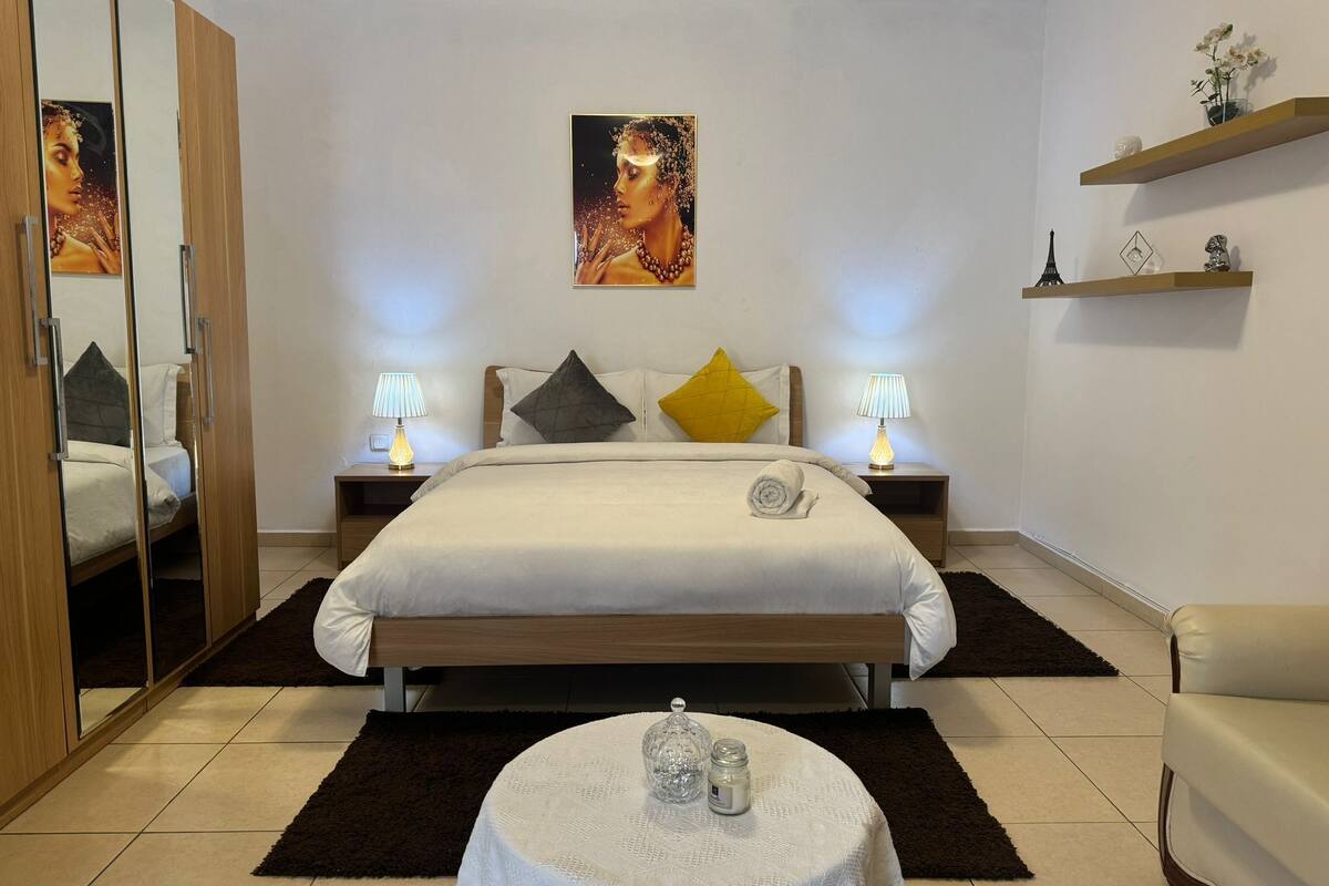 A bedroom with a bed, two bedside lamps, a round table with a decorative item, a mirror, and a painting on the wall.