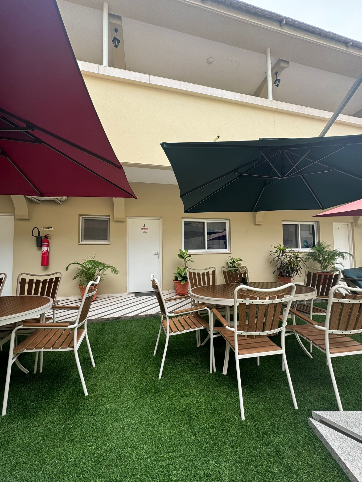 An outdoor patio area with round tables and chairs, potted plants, and umbrellas providing shade.