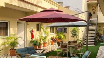 Un espace extérieur aménagé en terrasse, avec une table et des chaises, entouré de plantes en pot et agrémenté d’un parasol rouge.