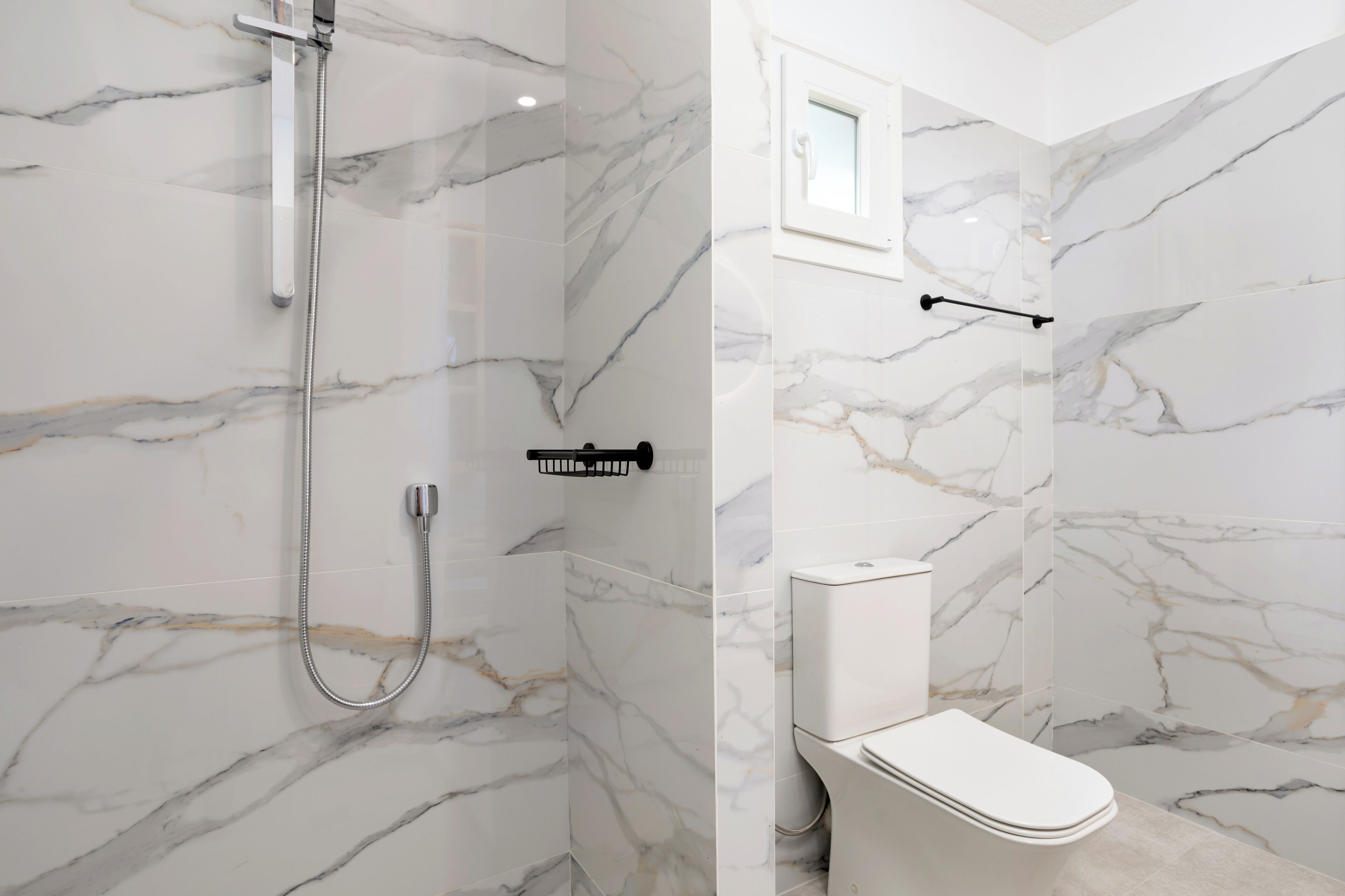Une salle de bain avec des murs en marbre, une douche, des toilettes et une petite fenêtre.