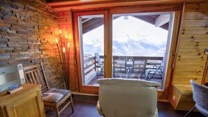 Una habitación acogedora con detalles de madera, un ventanal con vistas a la montaña, una silla de madera y una mesa con sillas afuera.