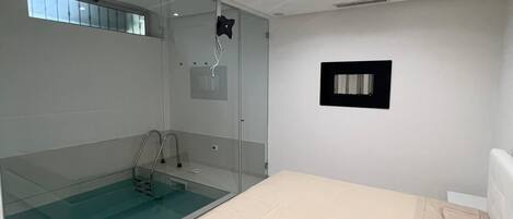 A modern bedroom with a glass-enclosed shower, a bed with a beige mattress, and a small black-framed picture on the wall.