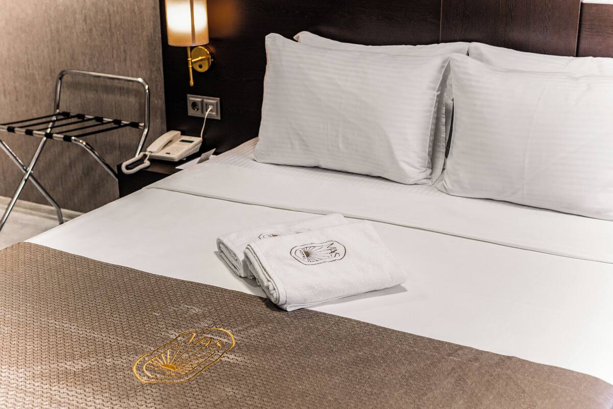 A neatly made bed with white linens and pillows, a folded towel with a logo, a metal drying rack, and a bedside telephone.