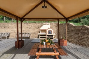 Une terrasse extérieure avec un mur en briques, des bancs en bois, une table dressée pour un repas et un four en briques.