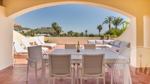 A terrace with a dining table and chairs, a sofa, and a view of palm trees and hills. - La Manga Club Los Olivos Apartment 600 (Atamaría)