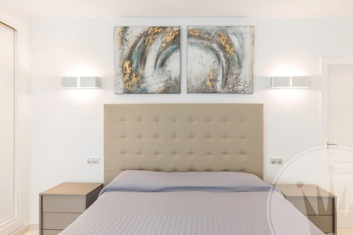 Une chambre à coucher avec une tête de lit capitonnée, un lit recouvert d’une couette grise, deux appliques murales et deux tableaux abstraits accrochés au mur.