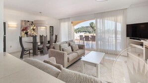 A modern living room with a sofa, dining table, and chairs, a television, and a view of the outdoors. - La Manga Club Los Olivos Apartment 600 (Atamaría)
