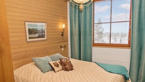 A bedroom with a wooden wall, a bed with a quilted bedspread, two pillows, a framed picture, a wall-mounted light, and a window with curtains.