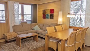 A cozy living room with a sofa, coffee table, and dining table with chairs. There are windows with blinds, a lamp, and a wall with colorful rugs.